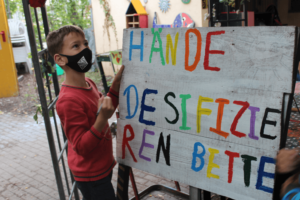 Kind mit Maske vor einem bunten Plakat auf dem Hände desinfizieren steht