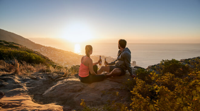 Paar genießt mit Erdinger Alkolfrei Zitrone den Sonnenuntergang auf einem Berg mit Blick über das Meer