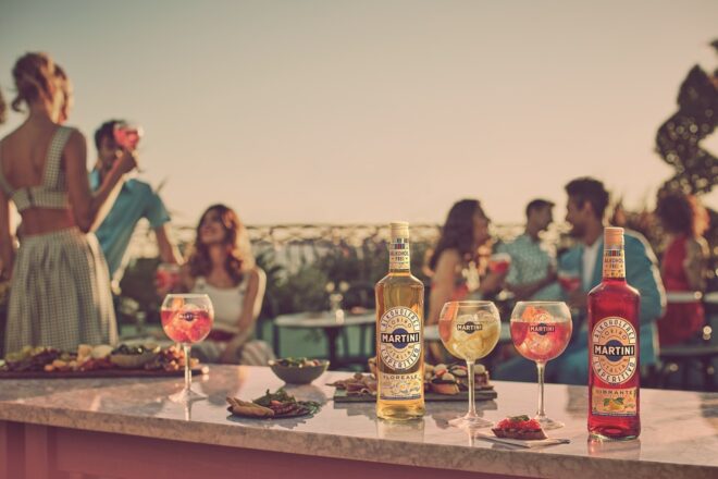MARTINI Alkoholfrei Flaschen und Gläser im Vordergrund und im Hintergrund Menschen die gemeinsam MARTINI genießen